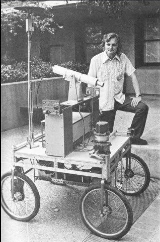 Hans Moravec standing beside the Stanford Cart, a four-wheeled camera robot, in the late 1970s.