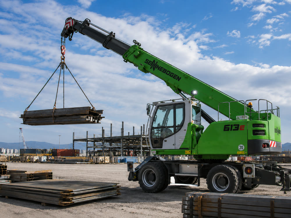 A pick-and-carry crane holds a bundle of steel sections on chains beside a steel-frame building.
